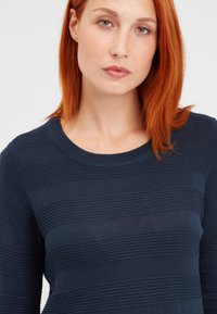 Granatowy sweter dzianinowy o poziomej fakturze prążkowanej, okrągłym dekolcie i długich rękawach. Materiał wydaje się miękki i lekko zwiewny.