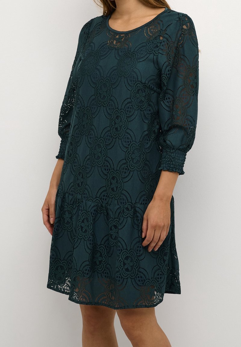 Femme portant une robe en dentelle vert foncé à motifs floraux, manches trois-quarts bouffantes et poignets froncés, debout devant un fond uni.