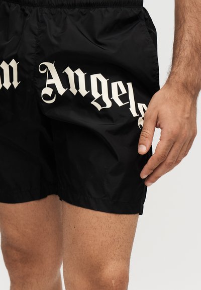 Μαύρο σορτς με λευκό γοτθικό κείμενο "Angels", που φοράει ένα άτομο που δείχνει το δεξί χέρι και το πάνω μέρος των μηρών πάνω σε ανοιχτό φόντο.