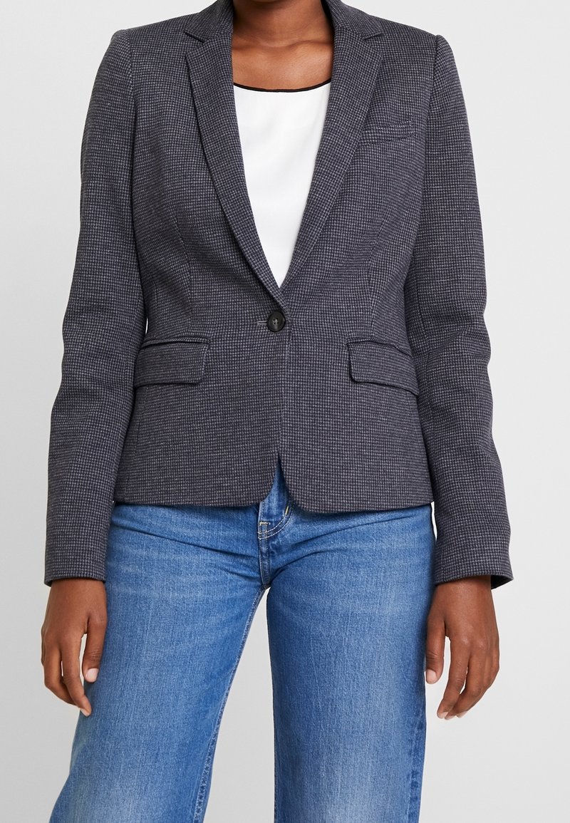 Personne portant un blazer ajusté à carreaux gris foncé par-dessus un haut blanc, associé à un jean bleu, debout devant un fond clair uni.