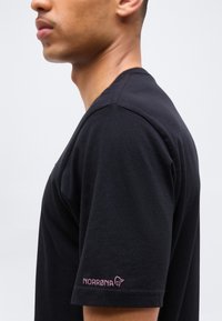 Άνδρας που φοράει μαύρο κοντομάνικο πουκάμισο με ροζ λογότυπο "NORRØNA" στο αριστερό μανίκι, σε απλό λευκό φόντο.