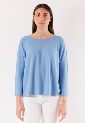 Donna con lunghi capelli castani che indossa una maglia a maniche lunghe azzurro chiaro e pantaloni bianchi, in piedi davanti a uno sfondo bianco uniforme.