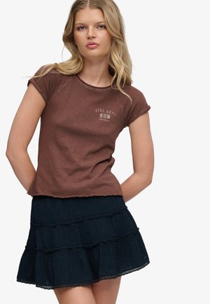 Blondynka w brązowej koszulce z krótkim rękawem z napisem "ATHL. DEPT 82 New York" oraz krótkiej, ciemnoniebieskiej sukience w warstwy.