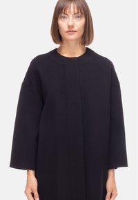 Cappotto oversize nero con scollatura rotonda, maniche lunghe e una texture liscia. Design minimalista senza chiusure visibili o motivi.