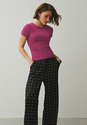 Woman wearing a fitted purple short-sleeve top and loose black pants with white polka dots, standing with hands in pockets.