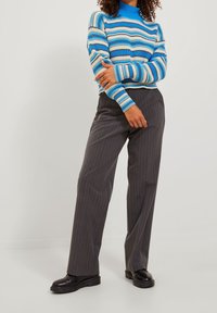 Gestreifter Pullover in Blau und Cremé mit hohem Kragen, kombiniert mit grauen Nadelstreifenhose und schwarzen klobigen Schuhen. Glatte Texturen und entspannter Schnitt.