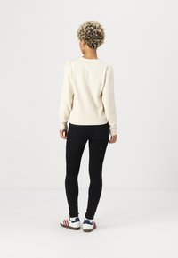 Krém színű, kötött crewneck pulóver, texturált mintával, hosszú ujjakkal, bordás szegéllyel és mandzsettákkal, fekete skinny farmernadrággal és fehér sportcipővel párosítva.