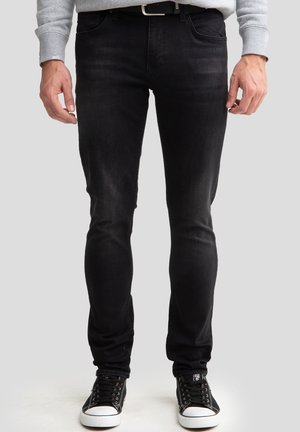 Vīrietis, tērpts melnās šaurās džinsu biksēs, melnās sporta kurpēs ar baltu zoli un pelēkā garroku krekliņā, stāv pret vienkāršu fonu.