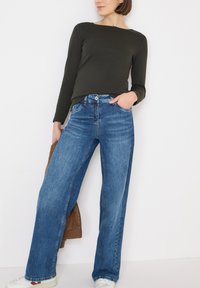 Donkergroen langarmshirt gecombineerd met wijde blauwe spijkerbroeken. De jeans hebben een klassiek ontwerp met vijf zakken en lichte vervaging. Witte sneakers.