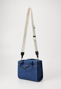 Sac en denim de forme rectangulaire, avec deux poignées supérieures et une bandoulière beige ajustable. Présente des accents en cuir noir et des coutures contrastées.