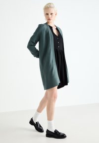 Long, open-front, green cardigan made of soft material, paired with a black dress, white socks, and black loafers, on a neutral background.