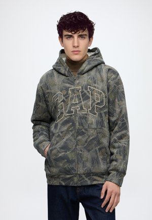 Jeune homme portant un sweat à capuche zippé camouflage vert et gris avec la capuche relevée et de grandes lettres GAP à l’avant, associé à un jean foncé.