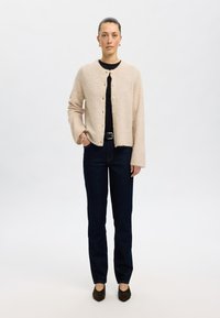 Beige knit cardigan with a round neckline, button closure, and loose fit, paired with dark blue straight-leg jeans and dark heels.