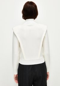 Witte gebreide trui met een hoge kraag, gestructureerde schouderdetails en een getailleerde taille. De achterkant heeft een gladde, ribbelige textuur.