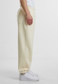 Lyse beige træningsbukser med en afslappet pasform, elastisk talje og en sidelomme, parret med hvide sneakers. Glat stoftekstur.
