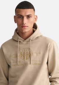 Felpa beige realizzata in morbido materiale, caratterizzata da un logo con design in rilievo. Presenta una tasca a marsupio frontale e un cappuccio con cordini.