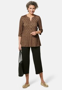 Braun gemusterte Tunika mit drei Viertelärmeln, Knopfleiste, kombiniert mit schwarzen, cropped Hosen, goldenen Slippern und einer schwarzen Tragetasche.