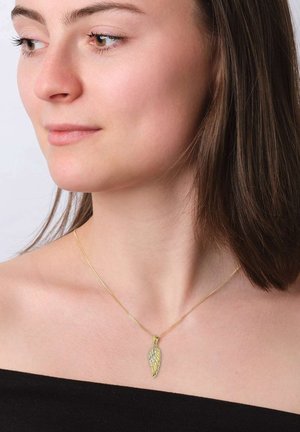 Junge Frau mit glattem braunem Haar, die eine goldene Halskette mit einem flügelartigen Anhänger trägt, der mit kleinen Steinen verziert ist.