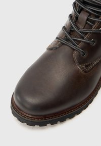 Bota de cuero marrón con puntera redondeada, cordones grises y costuras en contraste. Suela gruesa de goma con patrón de dibujo texturado.