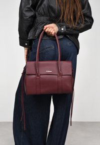 Bolso de mano de cuero burdeos con forma estructurada, doble asa y correa desmontable. Textura suave y detalle sutil del logo en la parte frontal.