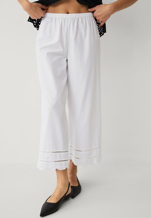 Kvinde iført hvide, brede cropped bukser med bølgekant og sorte spidse slingback-flats, stående mod en ensartet baggrund.