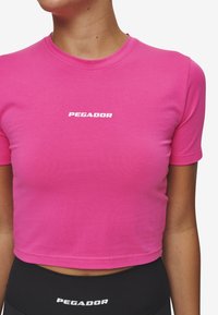 Roze cropped t-shirt met een ronde hals en korte mouwen. Het heeft een witte "PEGADOR" logo aan de voorkant. Gladde stof structuur.