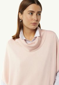 Hellrosa gestrickte Poncho mit weitem Kragen, strukturiertem Stoff und lockerer Passform, über einem weißen Hemd getragen. Runde Ohrringe sind sichtbar.