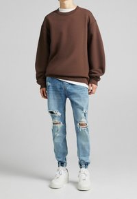 En person som bär en brun oversized sweatshirt, slitna ljusblå denimjeans och vita sneakers, står mot en enkel grå bakgrund.