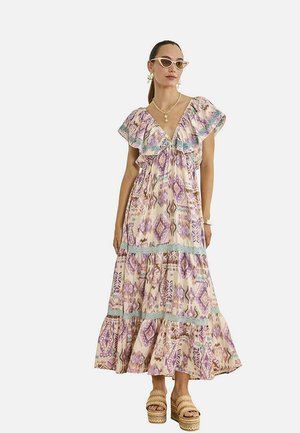 Mujer con un vestido largo morado de estampado con mangas con volantes, sandalias beige de plataforma, collar de perlas y gafas de sol estilo ojo de gato.