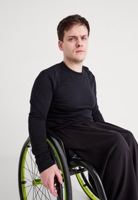 Fekete hosszú ujjú póló és laza fekete nadrág, amelyet egy kerekesszékes személy visel, zöld kiegészítőkkel ellátott kerekekkel és minimalista szürke háttérrel.