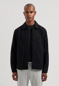 Svart teksturert skjortejakke med et markant hevet mønster, knappelukking og krage, kombinert med en svart T-skjorte og lysegrå bukser.