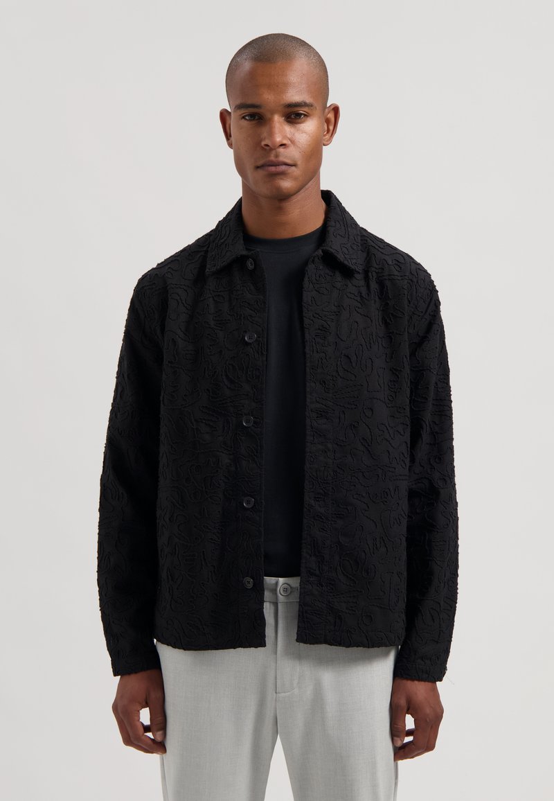Svart teksturert skjortejakke med et markant hevet mønster, knappelukking og krage, kombinert med en svart T-skjorte og lysegrå bukser.