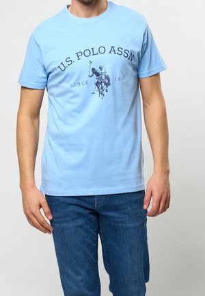 Homme portant un t-shirt bleu clair U.S. Polo Assn. avec un motif joueur de polo bleu foncé, associé à un jean bleu, se tenant devant un fond uni.