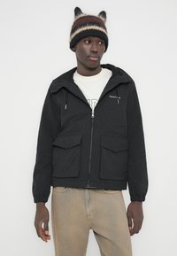 Μαύρο καρό τζάκετ με φερμουάρ και κουκούλα, με δύο τσέπες με καπάκι στο μπροστινό μέρος. Φορεμένο πάνω από ένα ανοιχτόχρωμο πουκάμισο και συνδυασμένο με μπεζ παντελόνι.