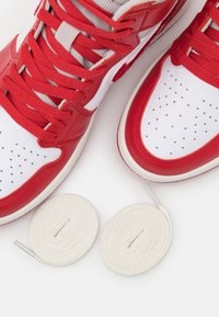 Červené a bílé vysoké tenisky s perforovanou špičkou, texturovanou červenou kůží a bílými tkaničkami. Vedle bot leží dva stočené bílé tkaničky.