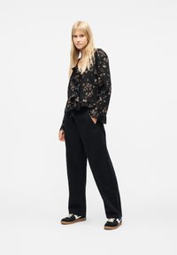 Νεαρή γυναίκα με μακριά ξανθά μαλλιά, που φοράει μαύρο μπλουζάκι με λουλουδάτο σχέδιο και βολάν, άνετο μαύρο παντελόνι και μαύρα αθλητικά παπούτσια με λευκές λεπτομέρειες.