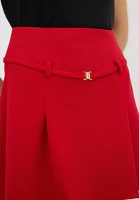 Falda roja con cintura ajustada, con una textura suave, un cinturón con hebilla dorada y una forma acampanada que cae a mitad del muslo.