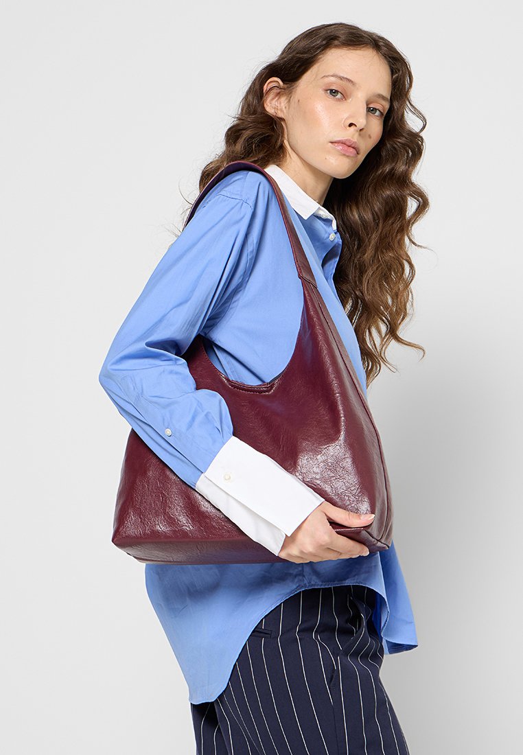 Bolso hobo de cuero burdeos con una textura suave, curvas suaves y una correa de hombro única, sostenido por una persona que lleva una camiseta azul.
