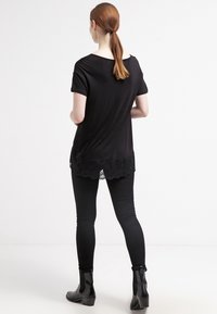 Kvinna med röd hår i hästsvans som bär en svart kortärmad topp med spetskant, svarta leggings och svarta ankelstövlar, stående vänd bort.