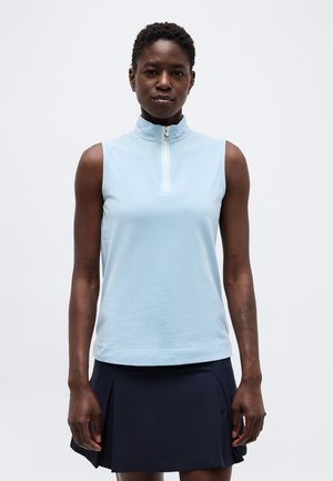 Vrouw die een mouwloos lichtblauw zip-top draagt en een zwarte geplooide rok, staand tegen een effen witte achtergrond.