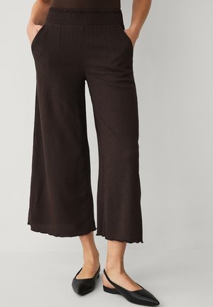 Kvinde iført løstsiddende, korte mørkebrune bukser med elastisk talje og sorte slingback flade sko, stående med hænderne i lommerne.