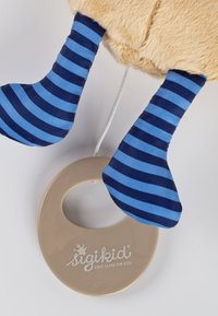 Das Plüschspielzeug verfügt über beiges Fell, blaue und marineblaue gestreifte Stoffbeine und einen hellbeigen Kunststoffring mit "sigikid"-Branding.