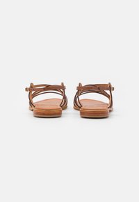 Sandalias de cuero marrón con correas cruzadas, parte trasera abierta y suelas planas. El diseño presenta un acabado suave y una correa ajustable en el tobillo.