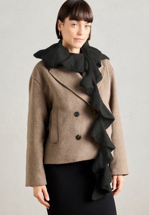 Femme aux cheveux courts bruns portant un manteau beige à double boutonnage et une longue écharpe noire à volants sur un fond gris uni.