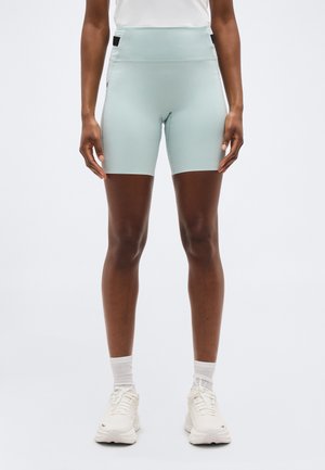 Κλείσιμο σε πρώτο πλάνο ενός ατόμου που φοράει ανοιχτό μπλε αθλητικό σορτς με ψηλή μέση, λευκές κάλτσες και λευκά παπούτσια τρεξίματος, στέκεται μπροστά σε απλό φόντο.