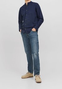 En man klädd i en mörkblå skjorta med knappar, blå jeans och beige mockaskor, som står med händerna i fickorna mot en grå bakgrund.