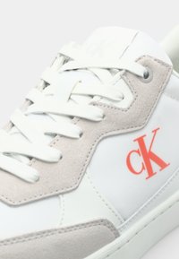 Bijele kožne tenisice s bež suknjenim naglascima, sa bijelim vezicama i neonsko narančastim "CK" logom sa strane.