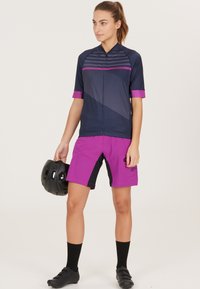 Cykeloutfit bestående av en marinblå kortärmad tröja med rosa detaljer och grafisk design, tillsammans med lila shorts och en svart hjälm.