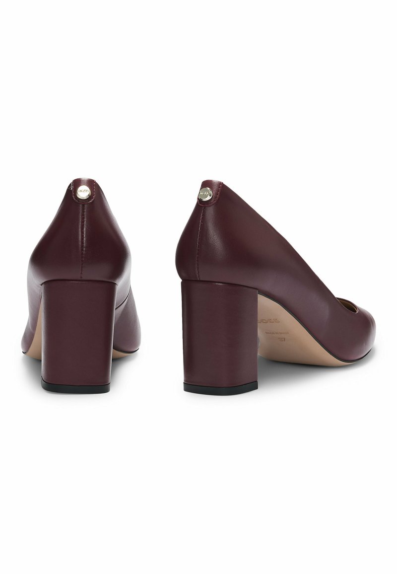 Scarpe con tacco alto in pelle bordeaux, con punta quadrata, tacco a blocco prominente e logo metallico sul retro. Texture liscia.