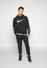 Felpa nera Nike con logo bianco, caratterizzata da maniche lunghe e tasca frontale. Pantaloni jogger neri con strisce laterali, abbinati a sneakers nere.